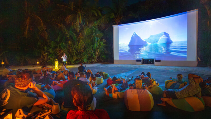 People sitting on bean bags outdoors, courtesy of Soneva and the Salt Group, are watching a large screen displaying an iceberg in the ocean. The setting is lush with tropical plants, creating a relaxed and immersive atmosphere under the night sky.