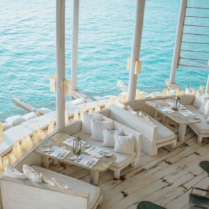 An open-air restaurant at the Maldives Luxury Resort features white wooden furniture and cozy cushioned seating, situated over serene, turquoise waters. Tables are set with menus and silverware, offering a picturesque dining experience with stunning ocean views—a true Soneva food paradise.