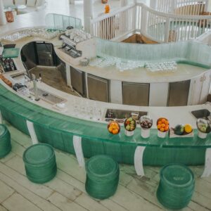 An aerial view of a modern, semi-circular bar with a green glass counter surrounded by matching stools at a Maldives luxury resort. The bar is equipped with various appliances, glassware, and a selection of fruits. The setting is bright and airy, with light wood accents and a spiral staircase.