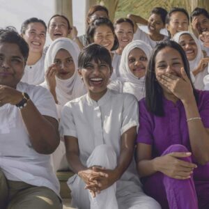 A group of women, some in white uniforms and hijabs and others in casual clothing, sit closely together, smiling and laughing. They appear to be outdoors under a sheltered area at a Maldives Charity event, enjoying each other's company.