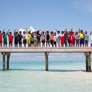 A group of children and adults stand on a wooden pier above the clear, turquoise waters of the Maldives. Everyone is wearing casual attire and waves at the camera. The sky is clear and sunny, and a sandy beach with greenery from Namoona is visible in the background.