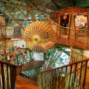 A vibrant and colorful interior of a treehouse features wooden spiral staircases, a large woven sphere centerpiece suspended from the ceiling, children’s artwork displayed on the walls, and multicolored triangular bunting hanging overhead—a Thailand exclusive experience by Soneva.