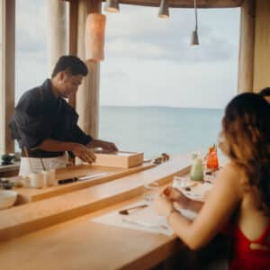 A chef, embodying the excellence of a Soneva Career, prepares food behind a wooden counter in a restaurant with large windows overlooking the ocean. Three people sit on the other side of the counter, watching the chef. The warm lighting creates a cozy atmosphere.