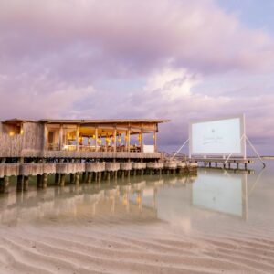 A scenic beachfront setup at a Maldives luxury resort, featuring a wooden structure on stilts over the water, illuminated warmly as evening sets in. Adjacent to it is an outdoor movie screen reflecting the serene, rippled water below. The sky is a pastel gradient of pink and purple clouds.