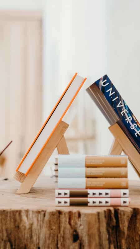 A wooden table displays various books. Two books are propped up on small wooden stands, including one titled "Universe." Additional books are stacked beside and in front of the stands. Softly lit shelves with more items, reminiscent of a serene Maldives holiday destination, are visible in the blurred background.