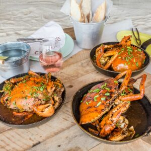 A round wooden table at the Maldives luxury resort Soneva holds three cast iron pans, each containing a cooked, sauce-covered crab garnished with green onions. Next to the crabs, there's a glass of pink wine, a bucket of bread, a small pail with utensils, and plates.