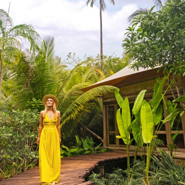 A woman in a yellow dress and straw hat stands on wooden steps, surrounded by lush tropical greenery. In front of a charming wooden cabin with large windows, tall palm trees rise against a partly cloudy sky in this exclusive Thailand experience brought to you by Soneva.