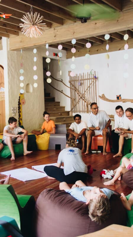 A group of children and adults participate in an interactive activity in a room decorated with colorful paper chains and hanging decorations, reminiscent of a family holiday in the Maldives. Some people are sitting on beanbags, while others are on the floor. Two adults appear to be leading the discussion.