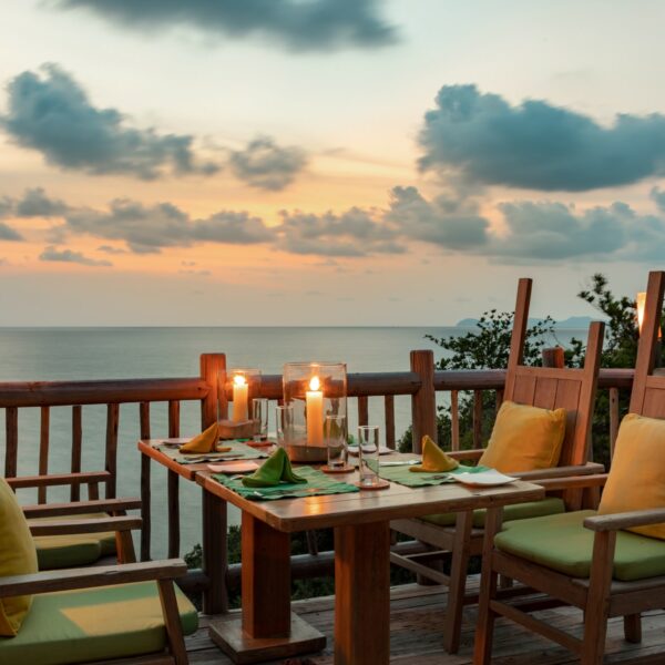 A cozy outdoor dining setting on a wooden deck overlooks the ocean at sunset, offering an exclusive experience in Thailand. The table is set with yellow cushioned chairs, green napkins, candles in glass holders, and tableware. Fluffy clouds dot the sky, with greenery visible on the right—a true Soneva escape.