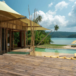 A serene tropical resort with a wooden deck and an infinity pool overlooking a calm beach. The deck has yellow cushions and a sizeable yellow umbrella. The scene includes lush greenery, palm trees, and a partly cloudy sky. A bamboo structure adds a rustic touch, part of the Soneva Exclusive Offers.