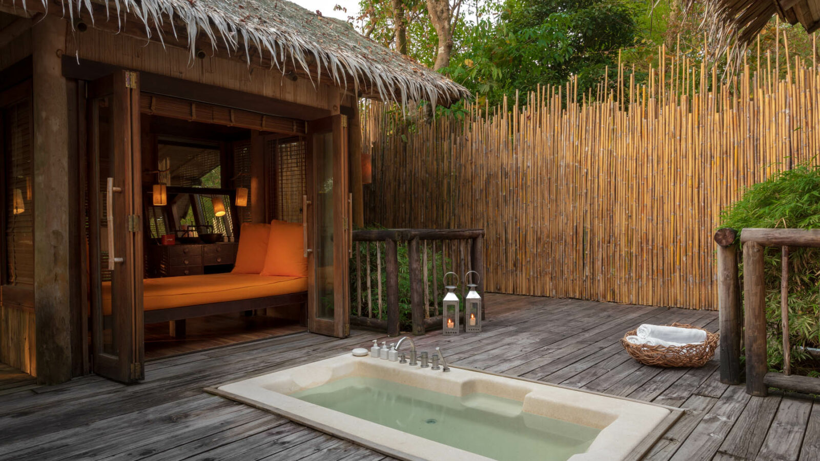 The image shows an outdoor wooden deck with a small Jacuzzi-style tub, perfect for a Thailand family holiday. To the left, there is a cozy cabin with a thatched roof and an orange sofa inside. The deck, reminiscent of Soneva resorts, is surrounded by a bamboo fence and adorned with lanterns and green plants.