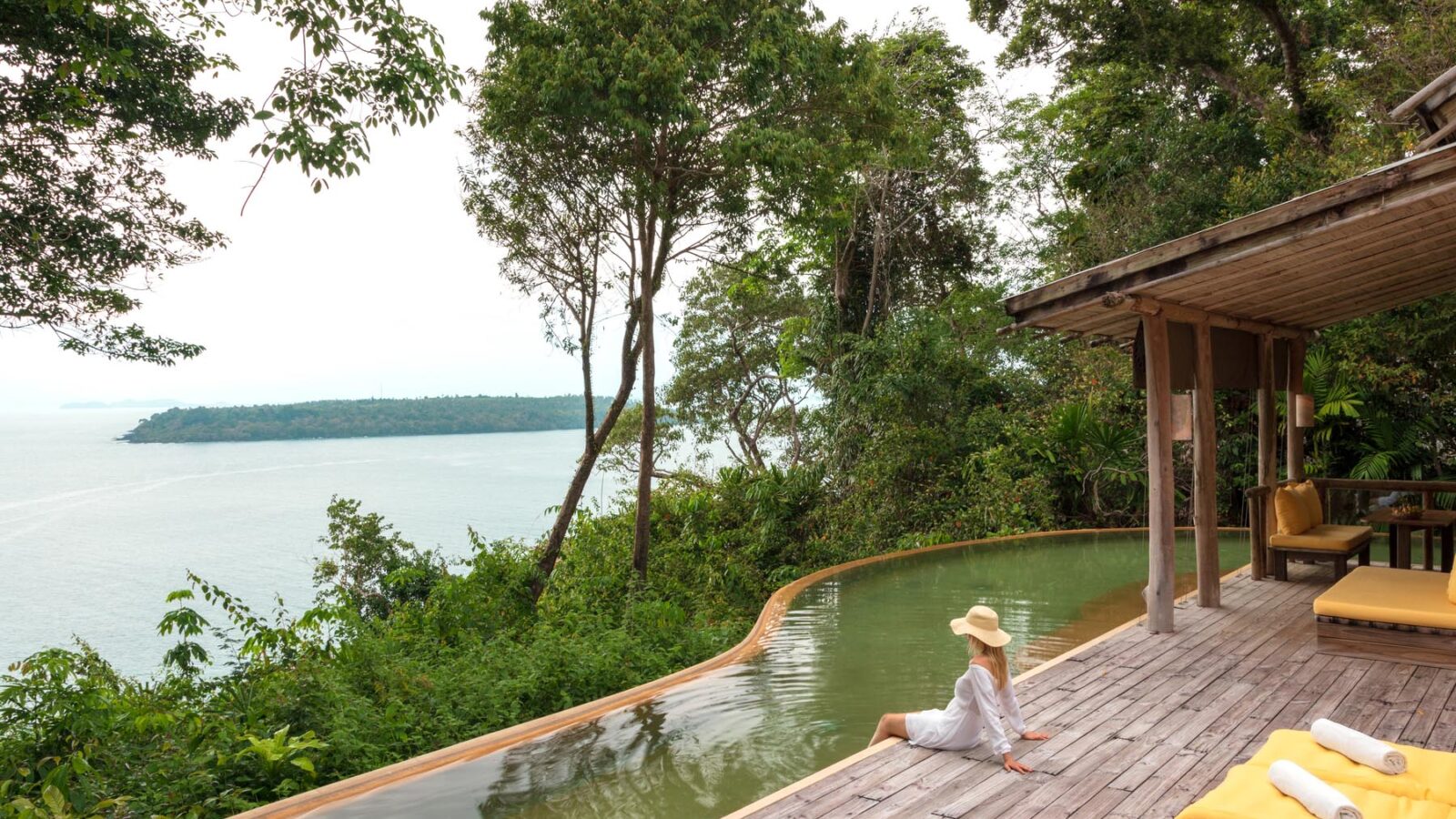 A woman in a white dress and hat sits at the edge of a curved, infinity pool surrounded by lush greenery, overlooking a vast body of water. Next to the pool is a wooden deck with yellow lounge chairs and a shaded seating area. The scene exudes relaxation and tranquility, perfect for a Thailand family holiday at Soneva.