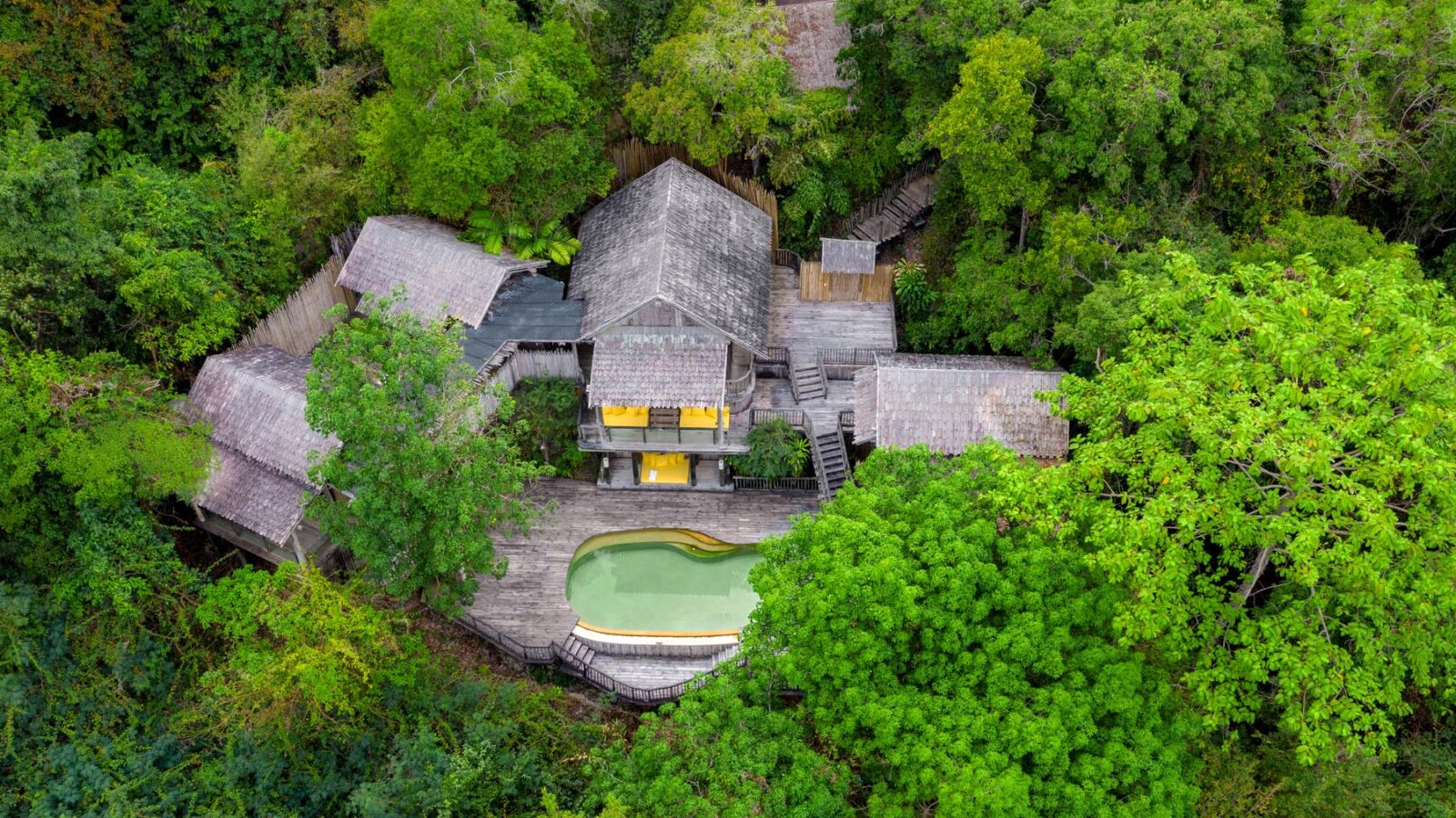 Aerial view of a luxurious, cabin-style house nestled in the dense, vibrant green forest of Thailand. The house has a large wooden deck with a swimming pool, surrounded by trees and foliage, creating a serene and secluded atmosphere perfect for a family holiday.