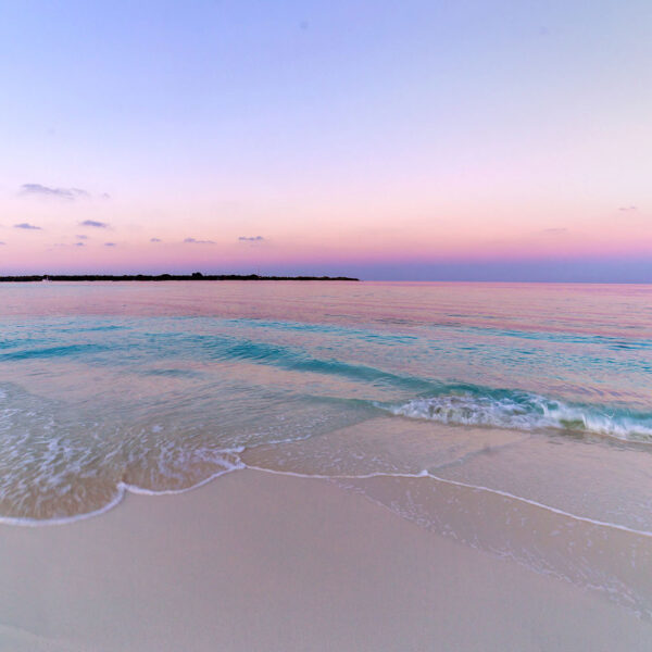 A serene beach scene at sunset, featuring gentle waves washing onto the sandy shore, embodies the essence of a Maldives exclusive experience. The sky is a gradient of soft pastel colors, transitioning from light pink to pale blue, with Soneva's distant strip of land visible on the horizon.