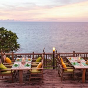 A wooden deck overlooking a calm ocean at sunset is set with two wooden dining tables, each surrounded by cushioned chairs in yellow and green hues. The tables are prepared with plates, glasses, and decorative plants, ready for an outdoor meal inspired by Soneva Exclusive Offers.