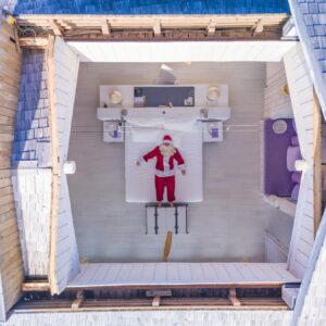 A person in a Santa Claus costume is lying on a bed in a bright, wooden, open-roof room at the Soneva resort in the Maldives. The room has light-colored wooden walls and floor, furnished with a desk, chair, and various household items. The open roof reveals a gray shingle exterior during this family holiday.