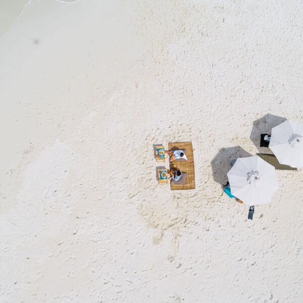 Aerial view of a sandy beach with two white umbrellas providing shade. Underneath the umbrellas are a couple of lounge chairs adjacent to each other and a woven mat. The beach, part of an exclusive Maldives experience, is mostly empty, with gentle waves washing up on the shore.