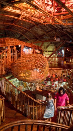 An intricate indoor space featuring a winding staircase made of bamboo and wood. The ceiling and walls are creatively designed with bamboo structures, offering an earthy and rustic ambiance. Two people, perhaps intrigued by Soneva Exclusive Offers, are seen walking down the stairs, exploring the unique environment.