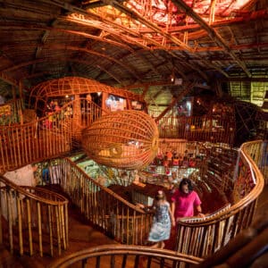 An intricate indoor space featuring a winding staircase made of bamboo and wood. The ceiling and walls are creatively designed with bamboo structures, offering an earthy and rustic ambiance. Two people, perhaps intrigued by Soneva Exclusive Offers, are seen walking down the stairs, exploring the unique environment.