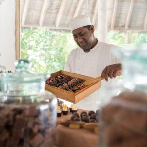 A chef wearing a white uniform and hat carefully holds a wooden tray filled with an assortment of chocolates. In the foreground, glass jars contain various types of chocolate pieces, blurred to draw focus on the chef, echoing the elegance found in Soneva Exclusive Offers.