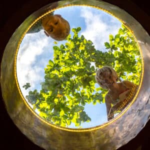 Two children look down through a circular glass window on their family holiday in the Maldives at Soneva. The sky is bright blue with fluffy clouds, and green leaves can be seen above them. The perspective is from below the window, capturing their curious expressions.