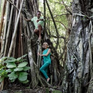 Two people, a man and a woman, are climbing and standing on the thick roots and trunk of a large tree in a dense forest. The man is higher up, holding onto a branch, while the woman stands lower, smiling and holding one of the tree's roots for support. It feels like their own Maldives Honeymoon Resort adventure.