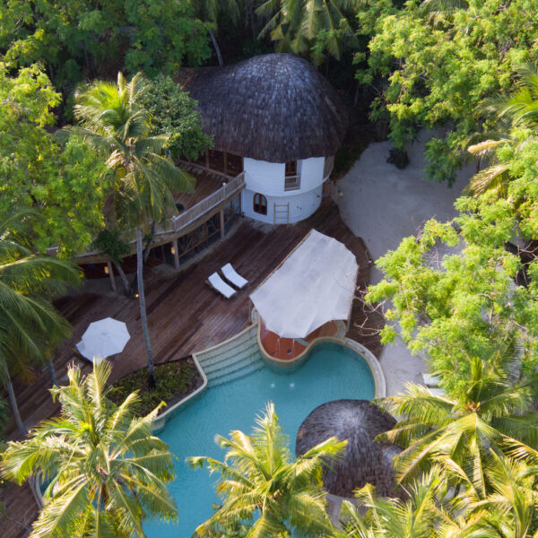 Aerial view of a Maldives Exclusive Resort featuring a two-story bungalow with a thatched roof surrounded by lush green palm trees. The Soneva bungalow overlooks a freeform swimming pool with a curved wooden deck, sun loungers, and a white canopy-covered seating area.