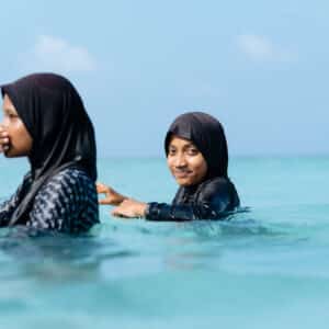 Two women wearing black burkinis swim in the clear, turquoise waters of the Maldives. One woman faces left, touching her face, while the other looks forward, smiling. The sky is clear and blue, adding a serene backdrop to this peaceful scene at Soneva Namoona.