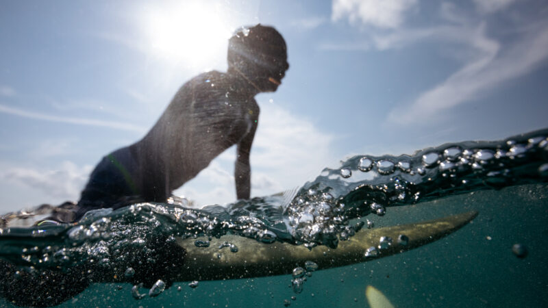 A person, silhouetted against a bright sun, rides a wave on a surfboard in the Maldives. The image is split to show both above and below the water, with bubbles and ripples in the foreground. This exclusive experience showcases a partly cloudy sky, epitomizing Soneva's unique offerings.