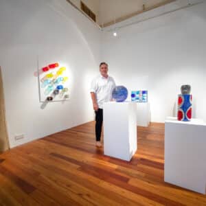 A man in a white shirt stands in an art gallery with wooden floors, surrounded by colorful abstract glass sculptures displayed on white pedestals and walls. The space, reminiscent of a Maldives Exclusive Experience at Soneva, is well-lit with a minimalist design.