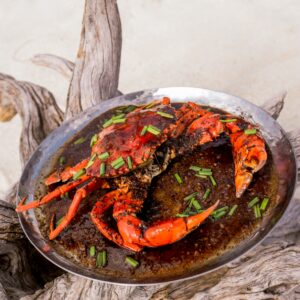 A silver plate holds a cooked, reddish-brown crab garnished with chopped green herbs. The plate is placed on a piece of driftwood, set against the sandy beach backdrop of a Maldives luxury resort.