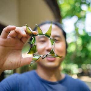 A person is holding up a circular piece of broken green and clear glass, shaped like an abstract star, in front of their face. The background is blurred, showing lush green trees and part of a building. Wearing a blue shirt, the scene captures the essence of a Maldives exclusive experience at Soneva.