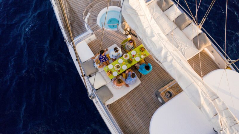 Aerial view of a group of people dining on a sailboat during their Maldives luxury yacht holiday. The boat's wooden deck is adorned with a yellow tablecloth under a sail. Plates and drinks are set on the table, surrounded by cushioned seating. The boat is in deep blue water.