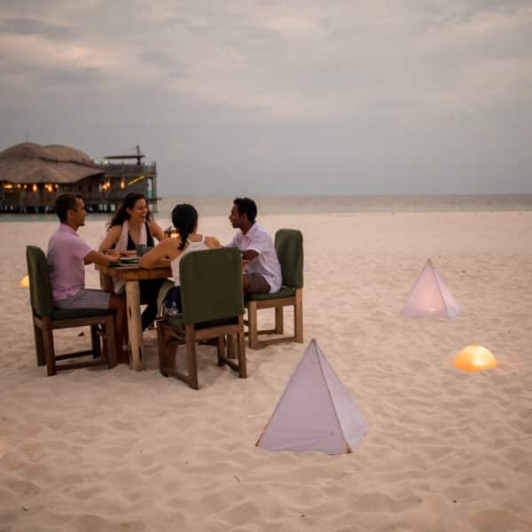 A group of four people are seated at a wooden table on a sandy beach in the Maldives, dining under an overcast sky. Ambient light emanates from pyramid-shaped lights surrounding them. In the background, overwater bungalows are visible, enhancing this exclusive Soneva experience.