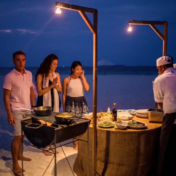 A group of people stands on a sandy beach in the evening, gathered around a table with food and a chef preparing a meal under two hanging lights. The atmosphere is casual, with attendees dressed in summer clothing, creating a relaxed and enjoyable beachside scene—a true Maldives Exclusive Experience at Soneva.