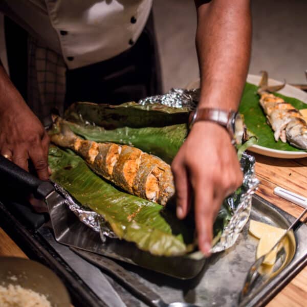 A person wearing a white shirt and apron is preparing grilled fish wrapped in green leaves, reminiscent of a Maldives exclusive experience at Soneva. The seasoned fish rests on a metal surface, with two cooked fish and lemon wedges on a nearby white plate. Cooking utensils and bowls are scattered on the wooden table.