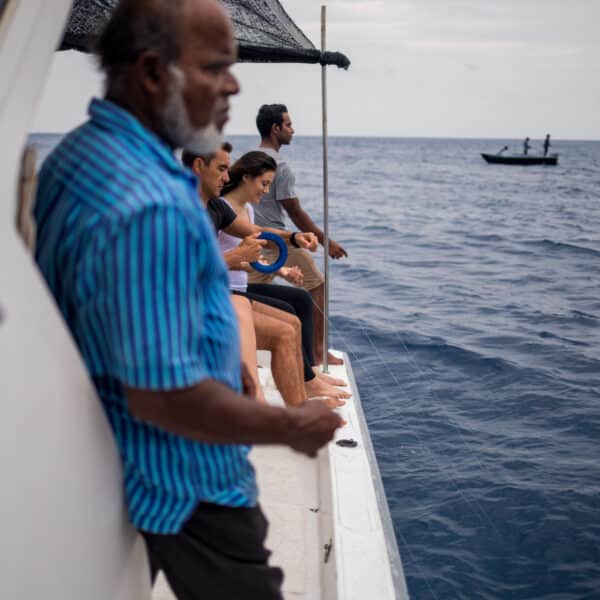 A group of people are on a fishing boat in the ocean, with three sitting at the edge and one person standing. Another small boat with two people is seen in the distance. The sky is overcast, and the water appears calm—a true Maldives exclusive experience akin to Soneva's serene offerings.