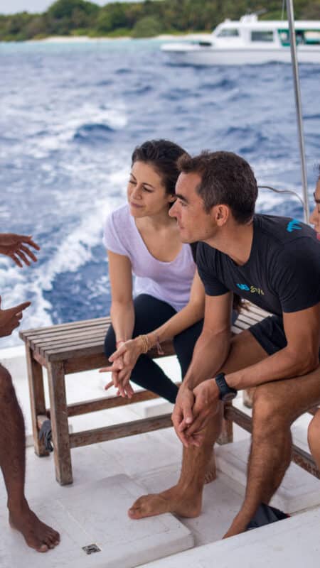 Four people are sitting barefoot on a small boat having a conversation. The boat is moving on the blue sea near an island with lush greenery. A white boat in the background complements this Maldives exclusive experience, making the scene appear casual and relaxed—a true Soneva moment.