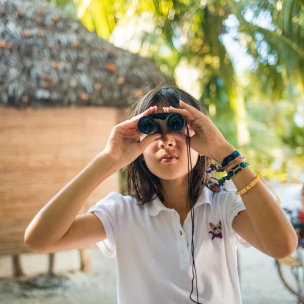 A young girl stands outdoors in front of a thatched hut, looking through binoculars. Clad in a white polo shirt and several colorful bracelets, she gazes around the picturesque setting. Palm trees and a bicycle dot the background, evoking an exclusive Soneva Maldives experience in this tropical paradise.