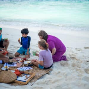 A small group of children and an adult are sitting on a mat on a sandy beach in the Maldives, having an exclusive picnic near the turquoise sea. There are various food items and drinks around them. The scene is bright and sunny, with the ocean and distant horizon in the background, epitomizing the Soneva experience.