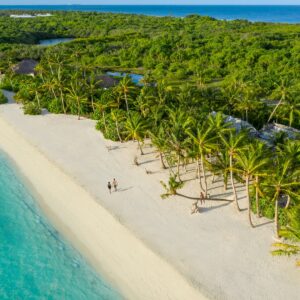 Aerial view of a tropical beach with clear turquoise water, white sand, and lush green palm trees. Two people are walking along the shoreline. Dense green vegetation covers the landscape in the background, extending towards the horizon with glimpses of the ocean at a Maldives luxury resort like Soneva.