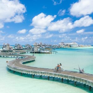 A scenic view of a tropical resort with winding wooden boardwalks over clear, turquoise water. Several thatched-roof huts are situated along the boardwalks. Two people ride bicycles on the boardwalk under a bright blue sky dotted with white clouds—discover it all with Soneva Exclusive Offers.