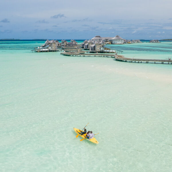 Aerial view of two people kayaking on clear turquoise waters towards overwater bungalows connected by wooden walkways. The scenery features a clear sky and vibrant blue tones of the ocean, giving a serene and tropical ambiance—truly a Maldives exclusive experience.