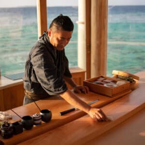 A chef dressed in traditional attire prepares sushi behind a wooden counter with the ocean visible through large windows in the background at a Maldives luxury resort. Various sushi ingredients and utensils are arranged on the counter, enhancing the calm and scenic ambiance of Soneva.