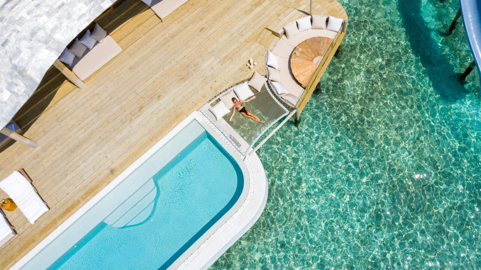 An aerial view of the Maldives luxury resort reveals a pool, a sunbathing deck, and clear turquoise water. At Soneva Fushi Villas, a person lies on a hammock suspended over the water, enjoying the sunny weather. The scene is serene and luxurious, with a mix of wooden and white structures.