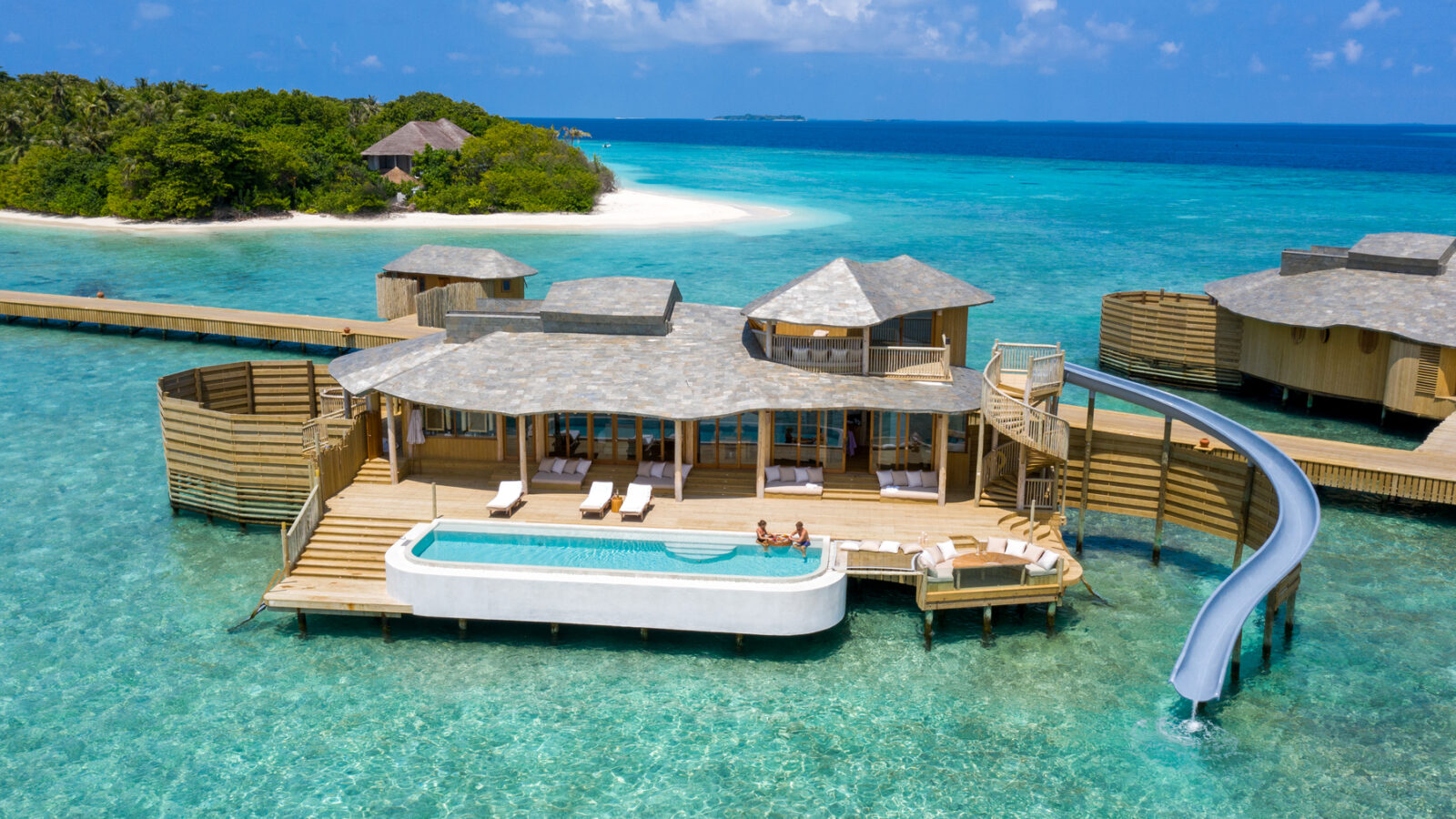 A luxurious overwater villa at the Maldives Luxury Resort, featuring a private pool and slide into the turquoise ocean, is shown. The Soneva Fushi Villas boast multiple sun loungers, shaded areas, and are connected to a wooden walkway. In the background, lush greenery and a white sandy beach are visible.