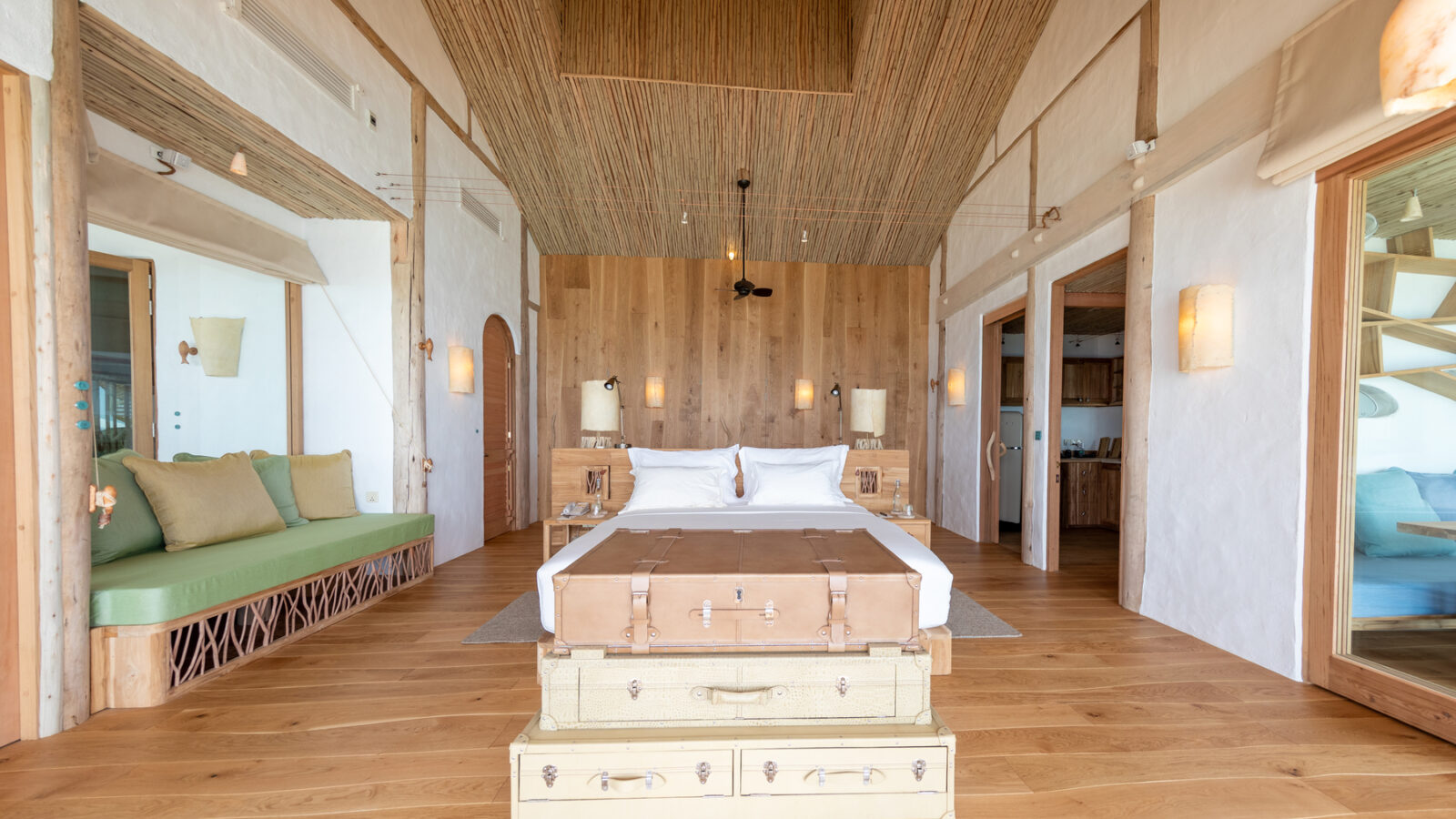 A spacious, rustic bedroom with a high thatched ceiling and wooden flooring at the Maldives Luxury Resort Soneva Fushi Villas. The bed, adorned with white linens, sits atop repurposed trunks. The room includes wooden accents, wall-mounted lights, a green cushioned bench, and large windows that let in natural light.