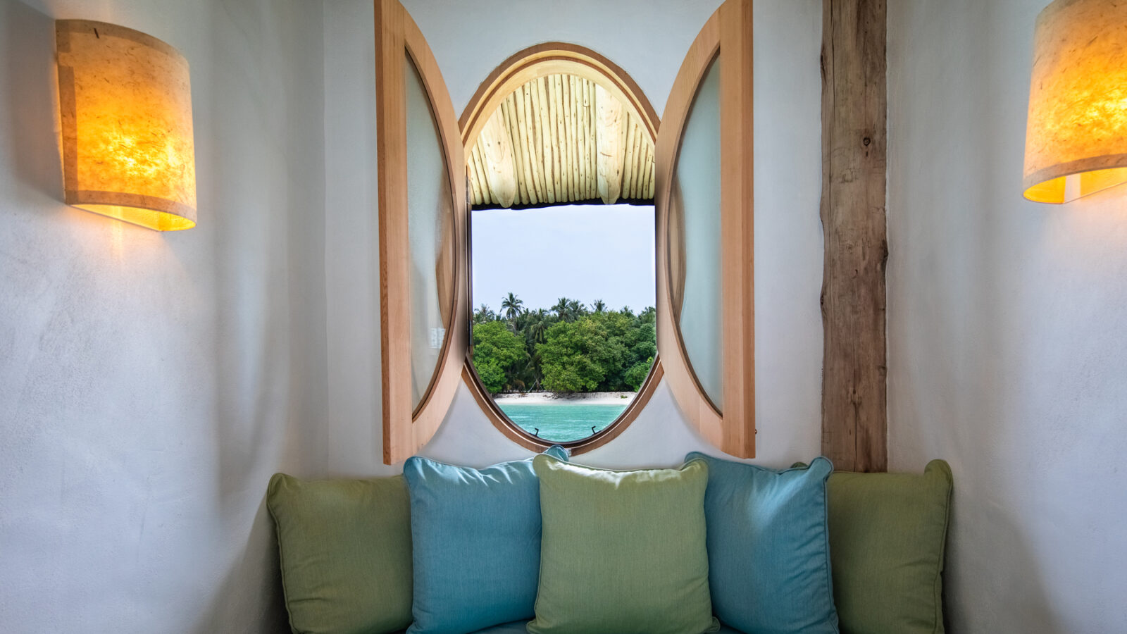 A cozy nook in one of the Soneva Fushi Villas features a cushioned seating area with blue and green pillows beneath a round wooden-framed window. The window offers a view of lush greenery and the ocean in the distance, reminiscent of a Maldives luxury resort. Two wall-mounted cylindrical lamps illuminate the space.