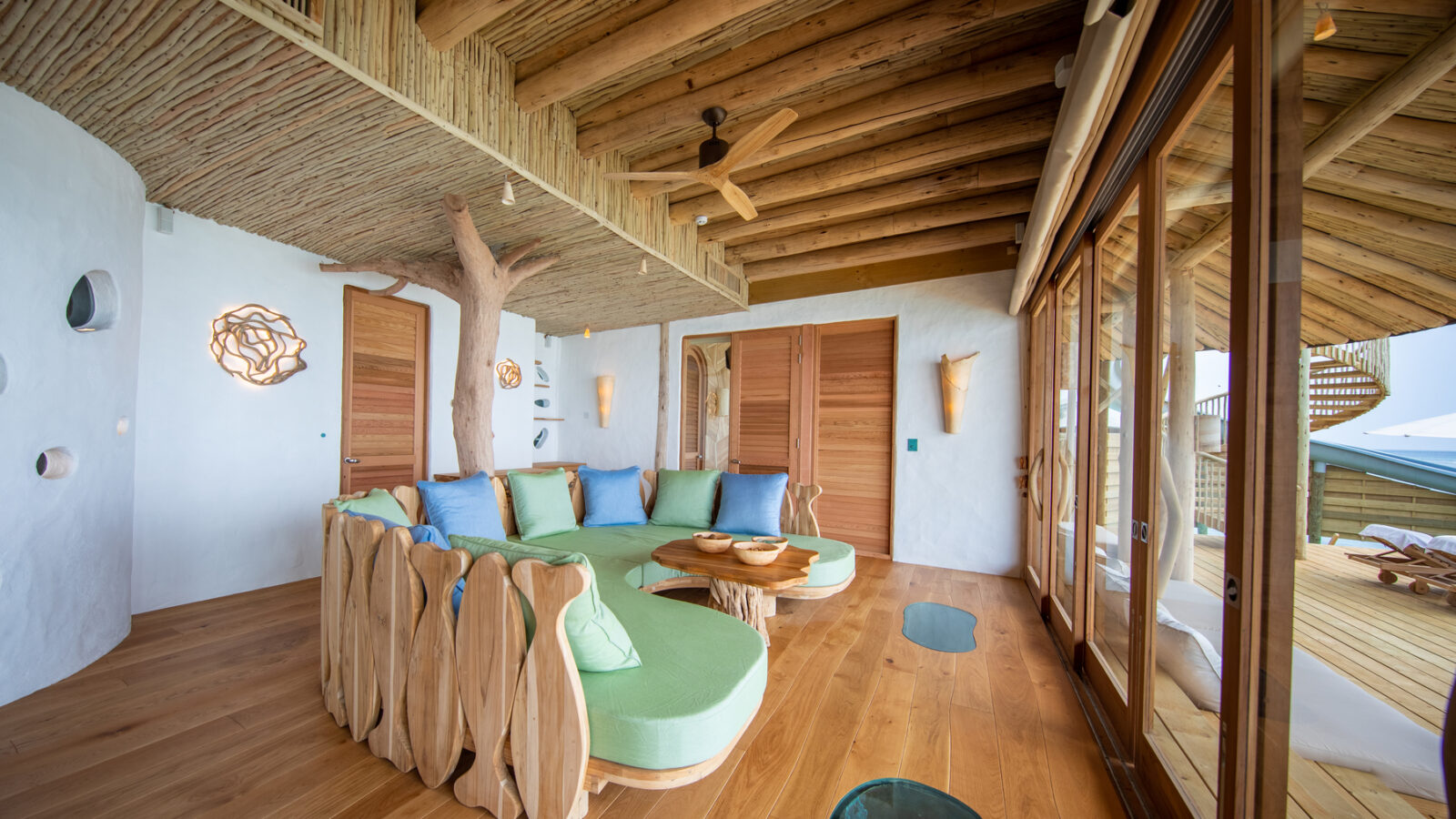 A bright, rustic living room with natural wooden elements, including a tree trunk pillar and a curved wooden sofa. The room features large glass doors opening to a deck with outdoor seating reminiscent of Maldives Luxury Resort vibes. Soft greens and blues add a touch of color to the minimalist space.