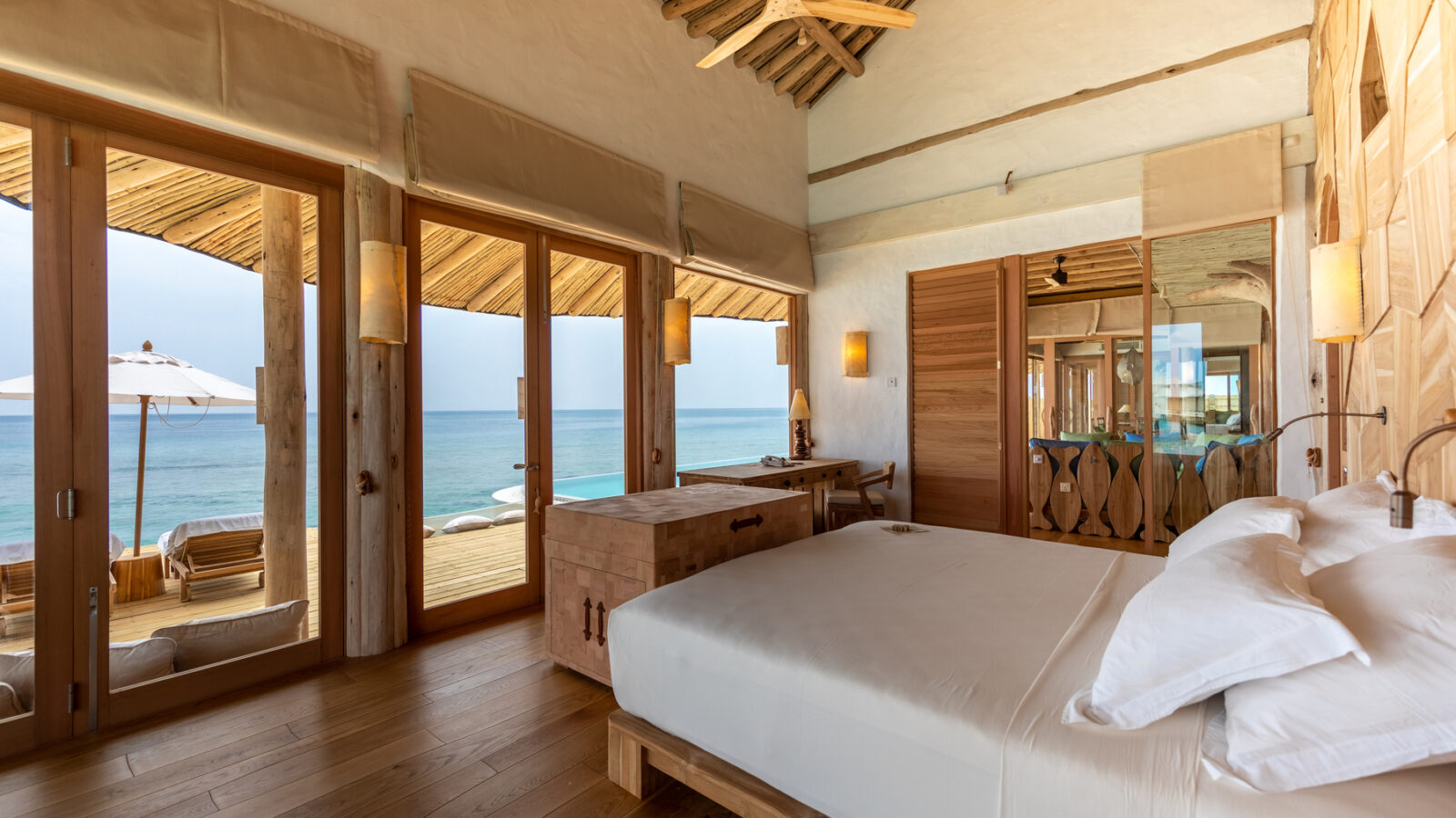 A spacious, rustic beachside bedroom at the Maldives Luxury Resort features wooden decor, a large bed, and floor-to-ceiling windows opening to a deck with lounge chairs and an umbrella. The room offers a stunning ocean view and a ceiling fan, creating a cozy, tranquil, and airy atmosphere typical of Soneva Fushi Villas.